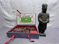 Wide shot showing a large standing terracotta warrior statue alongside an open decorative box containing four smaller warrior figurines arranged in red velvet compartments with fabric lid showing Chinese text.
