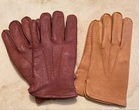 Two pairs of men's leather gloves laid flat on fabric surface, showing front side. Left pair is reddish brown with decorative stitching, right pair is tan with simpler finish.