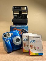 Photo showing front view of Polaroid 300 blue instant camera, Polaroid OneStep vintage camera on top of its box, and four packs of Polaroid 300 film in front.