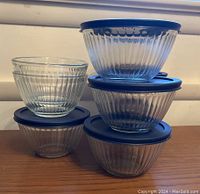Stack of nine ribbed clear Pyrex glass bowls, seven with blue round lids on top and two bowls without lids stacked on the side.