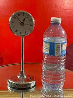 Front view of the silver-tone desk clock next to a water bottle for size comparison, showing the round clock face with Roman numerals and the flexible neck attached to the weighted base.