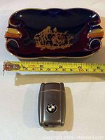 Full view of dark blue Limoges ashtray with gold figure imagery and measurements, resting above a BMW branded silver-tone lighter.