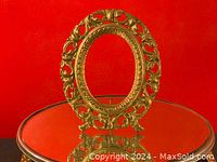 Full view of the ornate gold oval picture frame on a reflective surface against red background