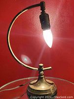 Side view of vintage brass arched arm half moon table lamp with pointed clear bulb lit, showing curved brass arm and round base.