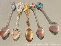 Top view of five vintage souvenir spoons laid out side by side, showing different ornate handles and colorful enamel emblems.