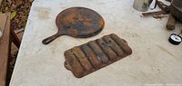 Round cast iron skillet and six-indent corn stick tray mold placed on a table, showing rust and condition.