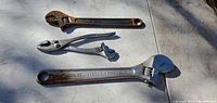 Photo showing three metal adjustable wrenches and plier combo tools on a metal surface with natural light, displaying condition and details.