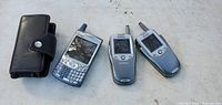 Photo showing three vintage Sprint cellphones: one black model with QWERTY keyboard and two silver flip phones with external antennas, along with a black leather Sprint carrying case with snap closure on the left.