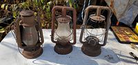 Three vintage metal lanterns displayed outdoors on a white table, two with glass and one without glass.