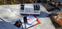 Photo showing a white electric Chef's Choice cutter sharpener with three sharpening slots, a black accessory, and instruction manuals placed on a white surface outdoors.