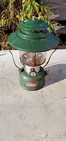 Vintage green Coleman lantern with glass globe showing signs of age and rust, photographed outdoors.