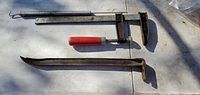 Photo shows a metal clamp with a red handle alongside a metal crowbar on a flat surface under daylight. Both items appear used with some surface rust.