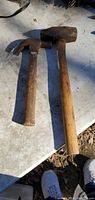 Photo showing a small claw hammer and a large mallet laid on a white surface outdoors with visible wear on wooden handles and some rust on the metal parts of tools.