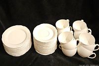 Overall view showing stacks of white plates, saucers, and matching cups with handles arranged on dark background.