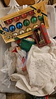 Box of vintage faceted Christmas tree light bulbs in multiple colors, with some Christmas textiles and ornaments in the bin