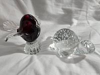 Two glass paperweights, a dark red bird with clear glass wings and a clear turtle with bubbles in the shell, placed on a white fabric background.