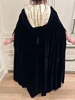 Back view of full-length black velvet cape with hood and visible cream silk lining, held by model
