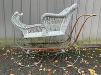 Side view of an antique white wicker baby pram showing metal frame, large spoke wheels, and wooden curved handle