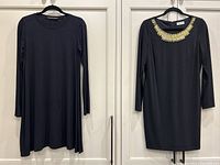 Two black dresses hanging on hangers against a white door, left dress plain and loose, right dress fitted with gold decoration at neckline