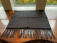 Full view of black scarf with fringed edge laid flat on wood table near window