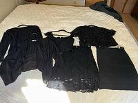 Seven black clothing items arranged on a bed: a sheer jacket, lace cropped top, lace skirt, bandage skirt, bodysuit, lace top, and sleeveless top.