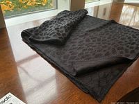 Black silky knit scarf or shawl folded on a wooden surface showing the subtle leopard pattern and knitted edges.
