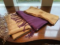 Three pashmina shawls folded and stacked on wooden surface near window, showing beige, purple with fringes, and tan/brown colors.