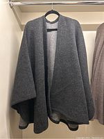 Front view of the dark grey knit cape hanging on a hanger. Shows the overall shape and color of the cape.