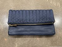 Front view of Deux Lux navy blue woven leather clutch with gold-tone zipper, showing fold-over flap design with woven leather and smooth leather back.