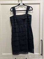Full view of black tiered pleated silk dress on hanger showing front side with wide straps and straight neckline.
