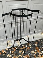 Front view of black wrought iron hanging pot rack with vertical rods and circular base support, photographed outdoors on ground with fallen leaves.