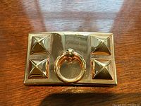 Front view of rectangular gold-tone buckle showing four pyramid studs and central ring pull