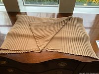 Beige knit scarf or shawl folded showing texture and edges on wooden surface with window background.