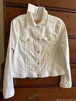Full front view of white denim jacket hanging on a wood dresser, showing collar, long sleeves, and brass buttons down the center and on chest pockets.