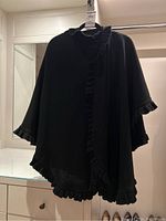 Front and side view of black ruffled edge wool poncho on hanger in a room with drawers and mirror, showing drape and ruffle details.