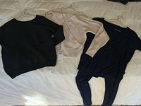 Three clothing items laid out on a bed: black knit sweater, beige knit sweater, navy blue lounge set with top and pants.