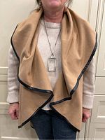 Front view of women wearing the camel-colored cape, highlighting the dark leather-like trim around the edges and the loose draping fit over a sweater and jeans.