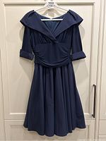 Full view of the navy blue dress hanging on a white hanger against a white cabinet door, showing the full skirt and wide collar.