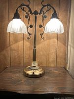Front view of vintage brass double light table lamp with white glass pleated shades and floral vine motif on arms and base on wooden surface.