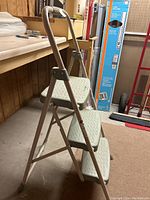 Side angle showing gray metal frame and three white textured plastic steps with oval pattern for grip. Safety handle at top is visible.