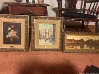 Three framed oil paintings side by side on carpet, one floral still life and two cityscapes with gold frames