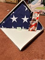 Folded American flag displayed in triangular white case with clear front on carpeted floor, showing star field prominently.