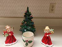 Full view of the Christmas tree, two red-dressed angel figurines, and the holly-decorated ceramic votive holder on a beige countertop.