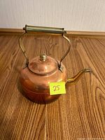 Copper pot with brass handle and knob on lid placed on wooden floor, labeled '25'