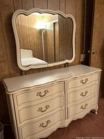 Cream colored French Provincial style dresser with six drawers and ornate handles, paired with a matching scalloped edge mirror mounted on the wall above.