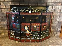 Front view of three-panel metal fireplace screen depicting a red holiday train carrying Santa, snowman, teddy bear, and other festive characters. Black background with gold stars and decorative gold scrollwork on top.