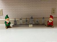Full view of Christmas gnome figurines (green and red) flanking the silver Christmas card holder with snowman and Christmas tree figures on a counter.