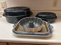 Image showing two black speckled oval enamelware roasting pans with lids, a silver metal bundt pan with dull gray interior, and three silver metal baking pans stacked inside each other.