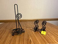 Paper towel holder on left with circular base and tall rod; napkin holder to right with symmetrical scroll pattern, on wooden surface