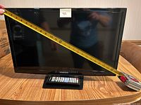 Samsung 24-inch flatscreen TV with measuring tape showing 24-inch diagonal screen size, and remote control in front on wooden table.